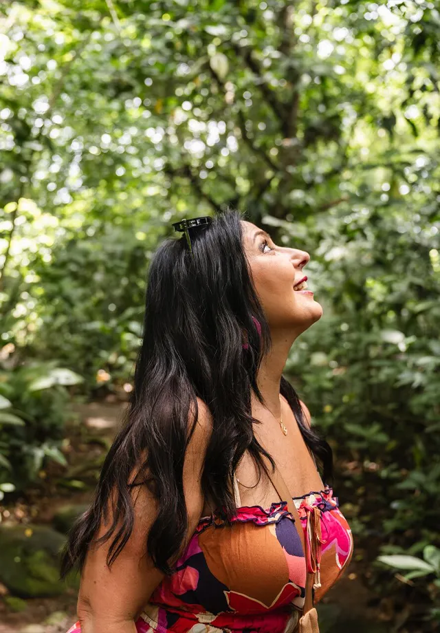 A woman with long dark hair in a floral dress smiles while looking upward in a lush green forest.